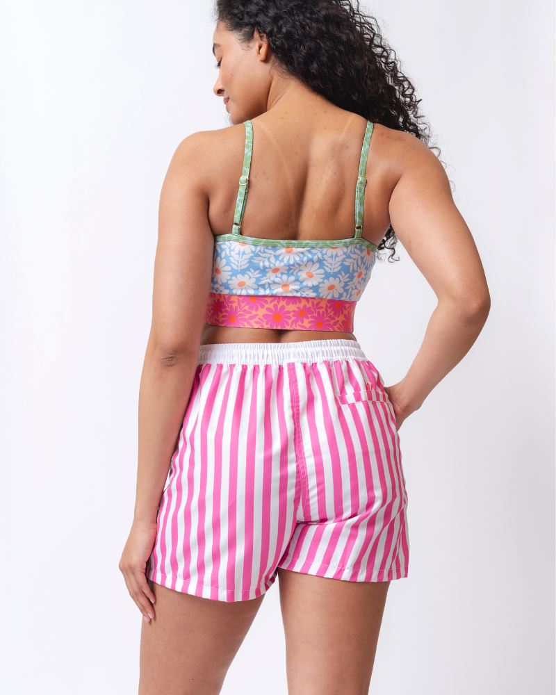 Photo of a woman wearing a pink and white striped board short swim bottom and a multi-colored floral swim bralette- back angle