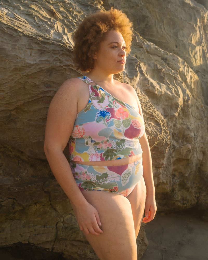 Photo of a woman posing on the beach wearing a colorful seashell inspired one-shoulder swim crop top and a colorful seashell inspired high waist swim bottom- side angle