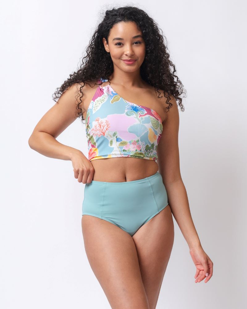 Photo of a woman wearing a light blue high waist swim bottom and a colorful seashell inspired one-shoulder swim crop top