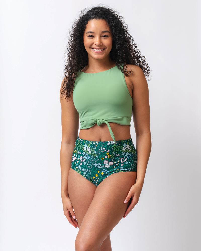 Photo of a woman wearing a light green knotted swim crop top and a dark green floral/ light green reversible swim bottom- floral side