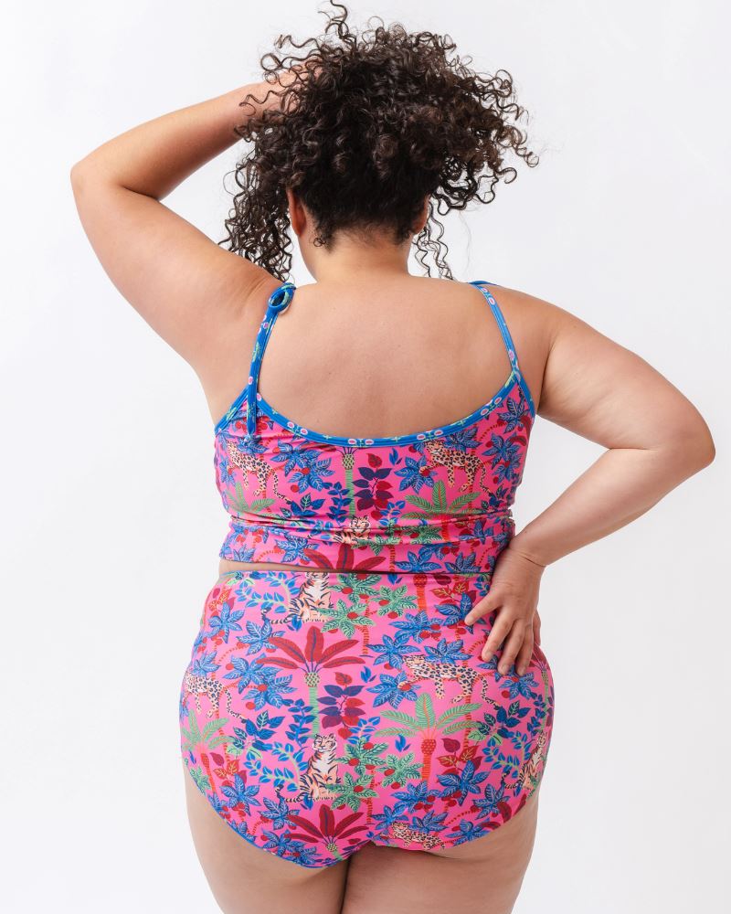 Photo of a woman wearing a bold pink and blue print featuring tigers and leopards shoulder-tie swim crop top and a bold pink print featuring tigers and leopards/ blue floral reversible swim bottom- tiger side- back angle