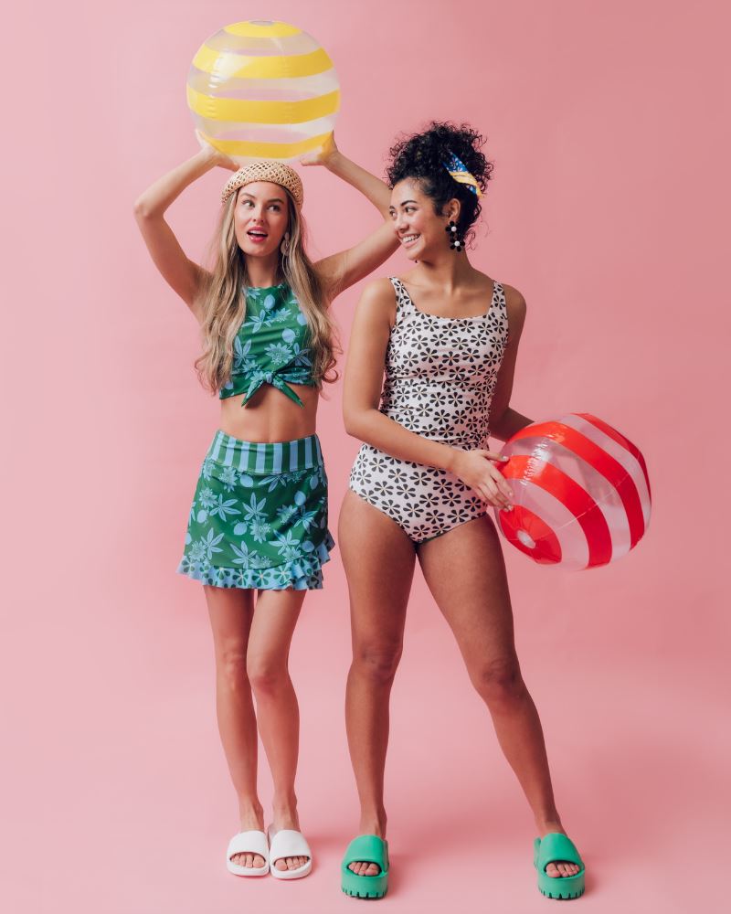 Photo of two women wearing a black and white floral square neck swim top and a black and white floral swim bottom and a green and blue floral swim crop top and a green and blue swim skirt bottom