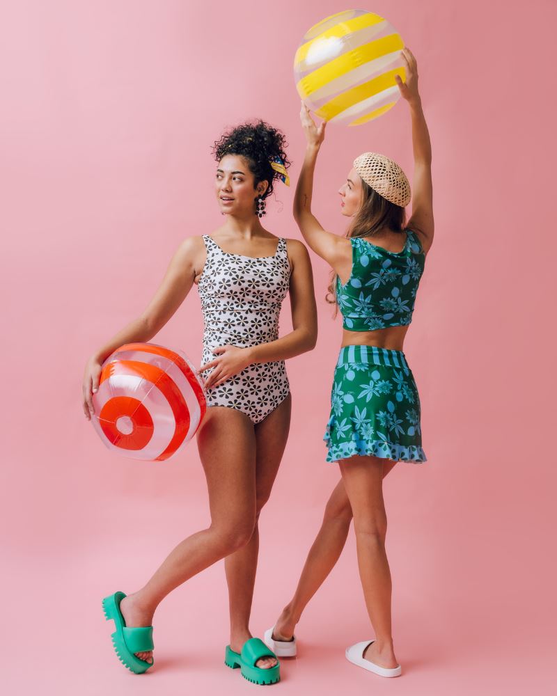Photo of two women wearing a black and white floral square neck swim top and a black and white floral swim bottom and a green and blue floral swim crop top and a green and blue swim skirt bottom