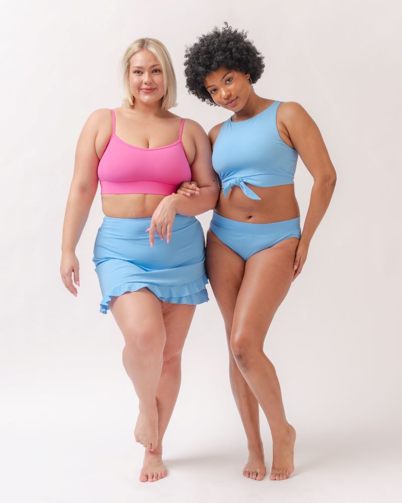 Photo of two women wearing a light periwinkle blue classic swim bottom with a light periwinkle blue swim crop top and a light periwinkle blue swim skirt bottom with a dark pink swim bralette