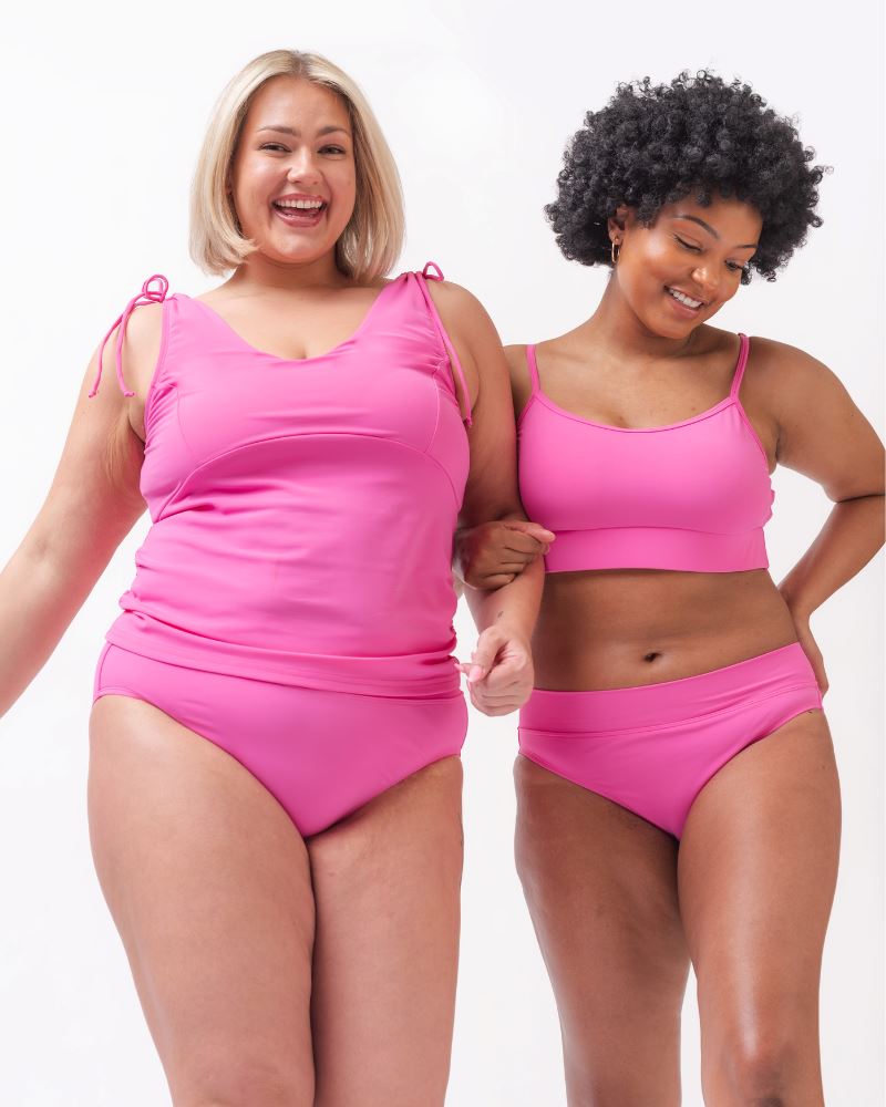 Photo of two women one with her hand on her hip wearing a pink swim tankini with pink low waist swim bottoms and the other wearing a pink cropped swim top with pink low waist swim bottoms