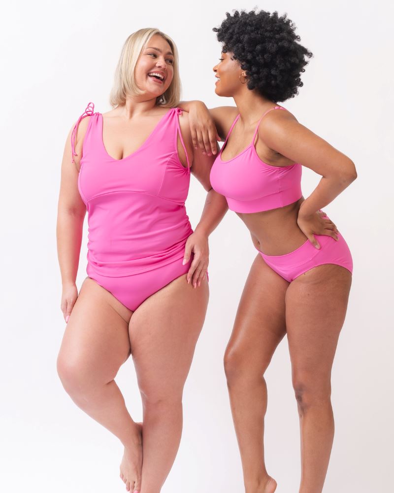 Photo of two women one with her hand on her hip wearing a pink swim tankini with pink low waist swim bottoms and the other wearing a pink cropped swim top with pink low waist swim bottoms