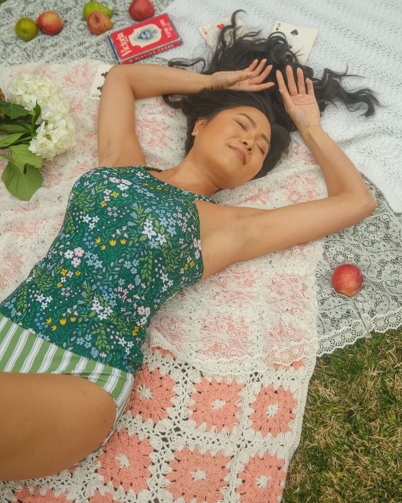 Photo of a woman laying down wearing a dark green floral double-cinch tankini swim top and a green and white striped/ pink and white floral reversible swim bottom- striped side