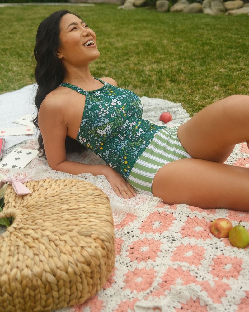 Photo of a woman laying down wearing a dark green floral double-cinch tankini swim top and a green and white striped/ pink and white floral reversible swim bottom- striped side- side angle