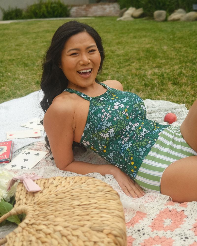 Photo of a woman laying down wearing a dark green floral double-cinch tankini swim top and a green and white stripe/ pink and white floral reversible swim bottom- striped side