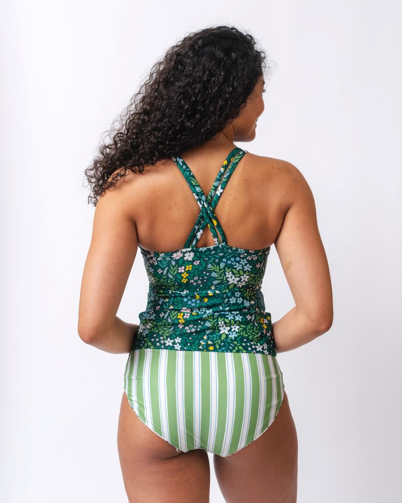 Photo of a woman wearing a dark green floral double-cinch tankini swim top and a green and white striped/ pink and white floral reversible swim bottom- striped side- back angle