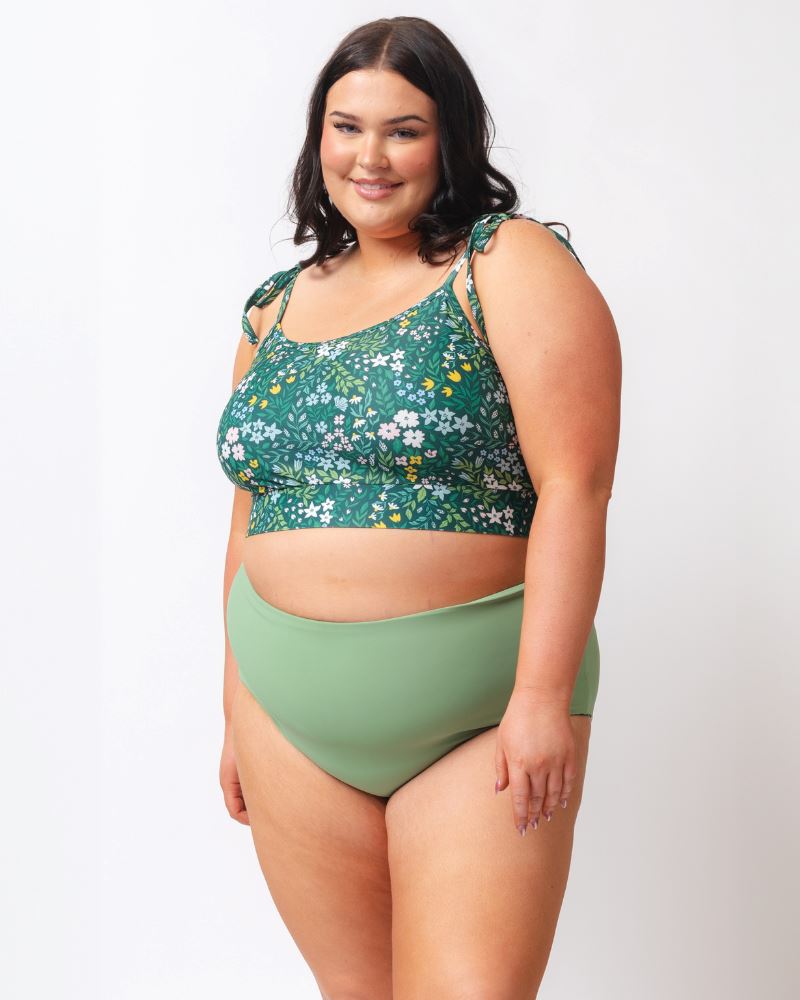 Photo of a woman wearing a dark green floral/ light green reversible swim bottom - light green side and a dark green floral shoulder-tie swim crop top
