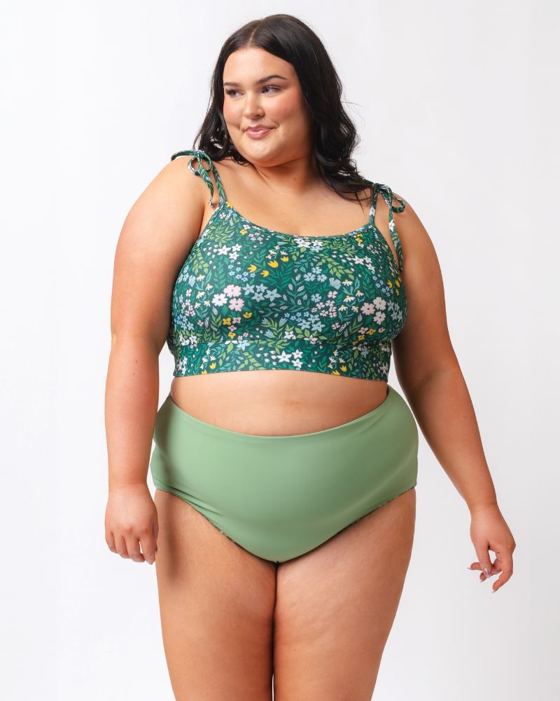 Photo of a woman wearing a dark green floral shoulder-tie swim crop top and a dark green floral/ light green reversible swim bottom- light green side