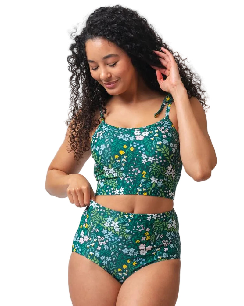 Photo of a woman wearing a dark green floral shoulder-tie swim crop top and a dark green flora/ light green reversible swim bottom- floral side