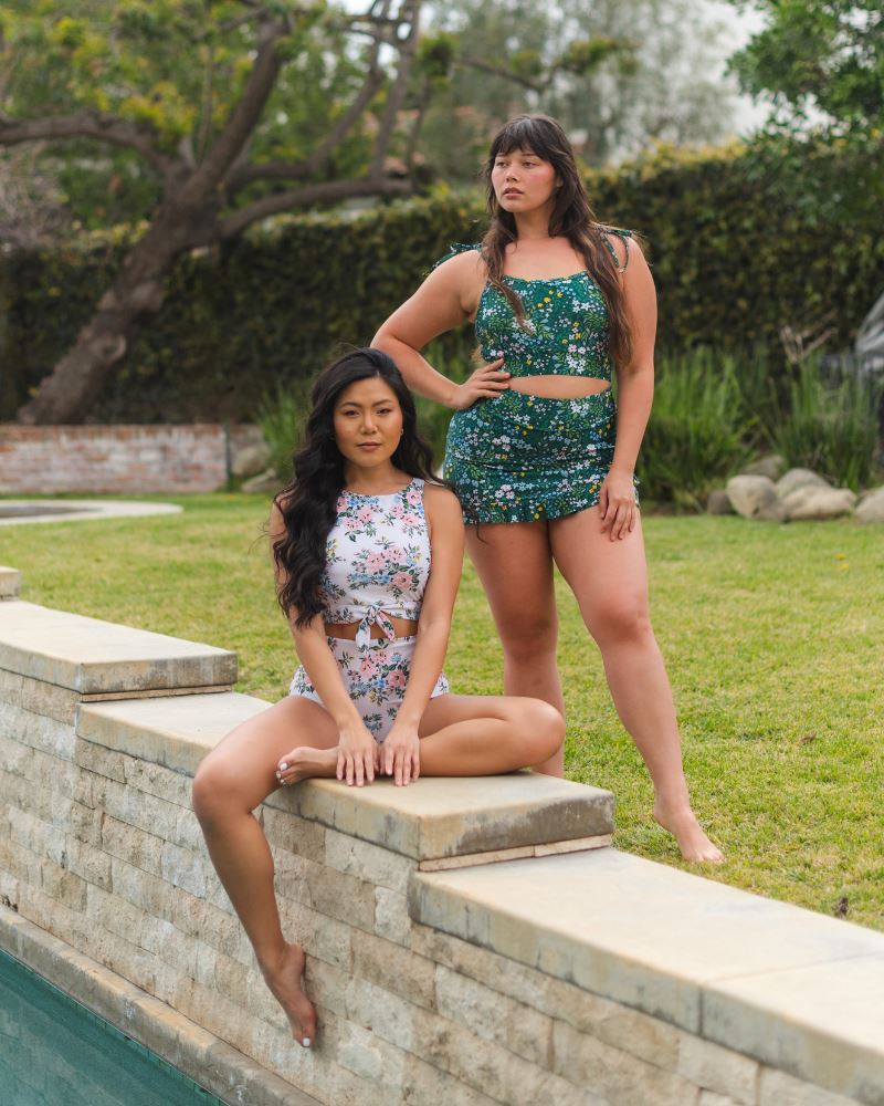 Photo of two women posing together wearing a dark green floral shoulder-tie swim crop top and a dark green floral swim skirt and the other woman wearing a pink and white floral knotted swim crop top and a floral swim bottom