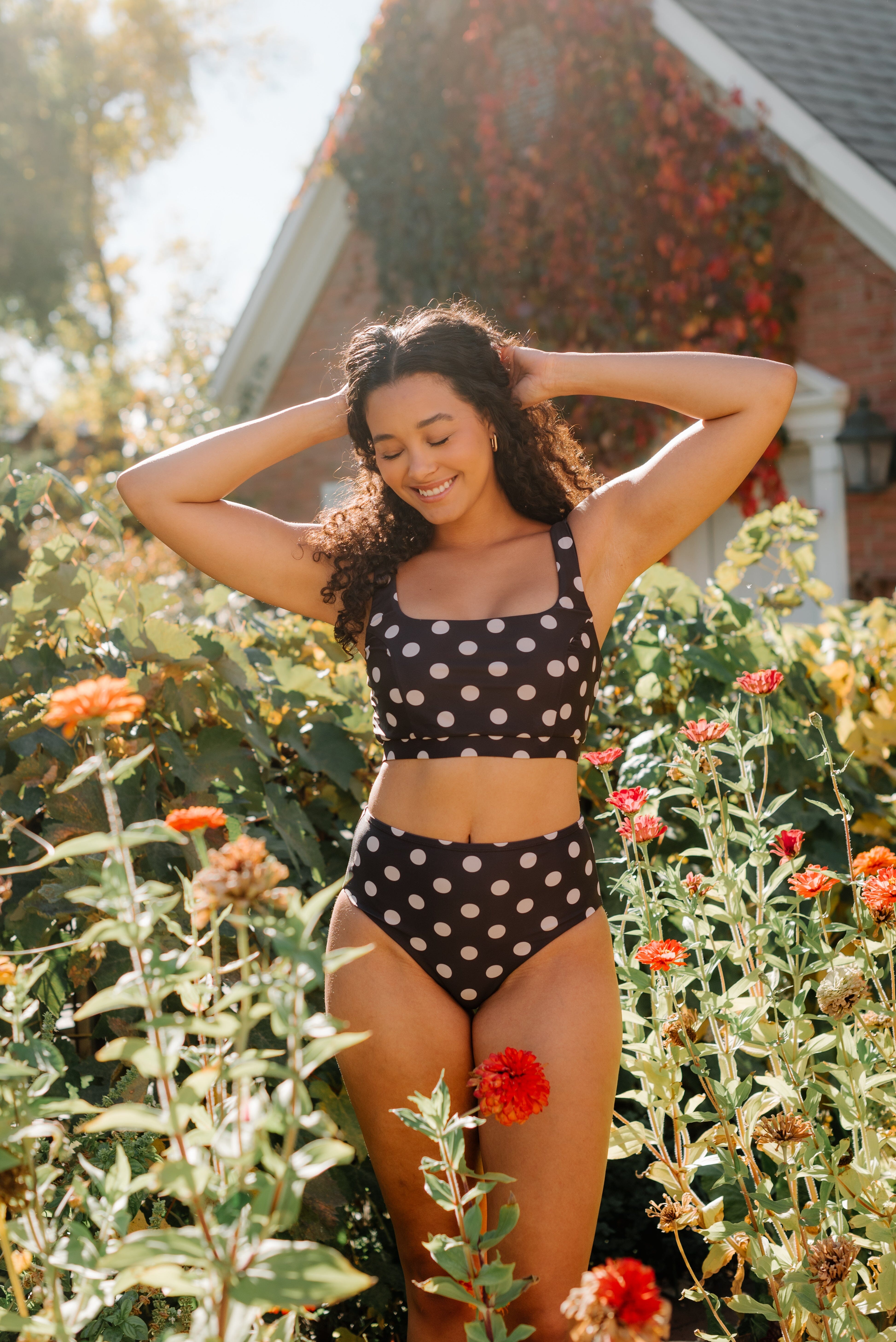 Woman in a polka dot swimsuit standing in a garden with flowers and a building in the background.