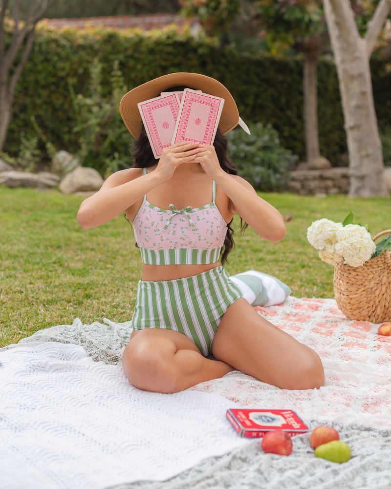 Photo of a woman posing outside wearing a pink and green floral swim bralette and a white and pink floral/ green striped reversible swim bottom- striped side