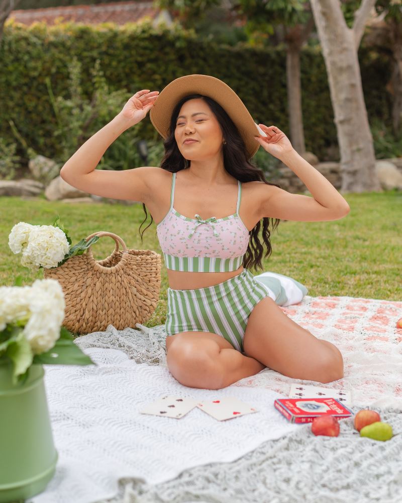 Photo of a woman posing outside wearing a pink and green floral swim bralette and a white and pink floral/ green striped reversible swim bottom- striped side