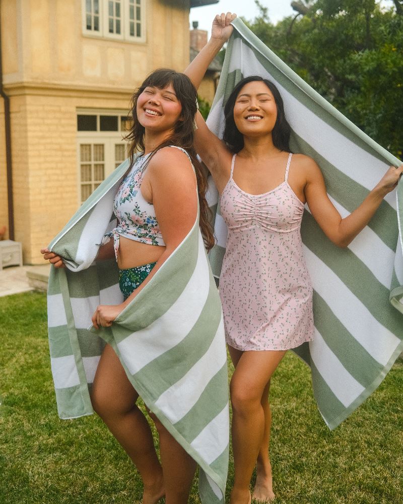 Photo of two women posing together wearing a pink floral swim dress and the other woman wearing a white and pink floral swim crop top and a dark green floral/ light green reversible swim bottom- floral side