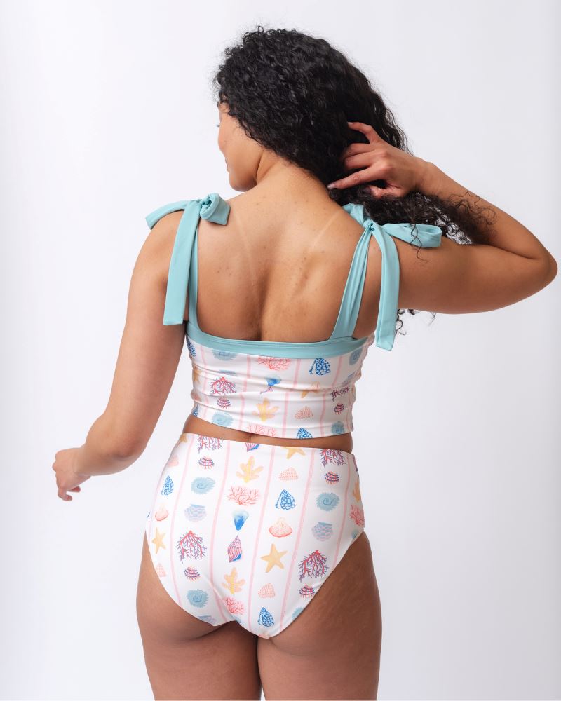 Photo of a woman wearing a seashell striped high waist swim bottom and a seashell striped shoulder-tie swim crop- back angle