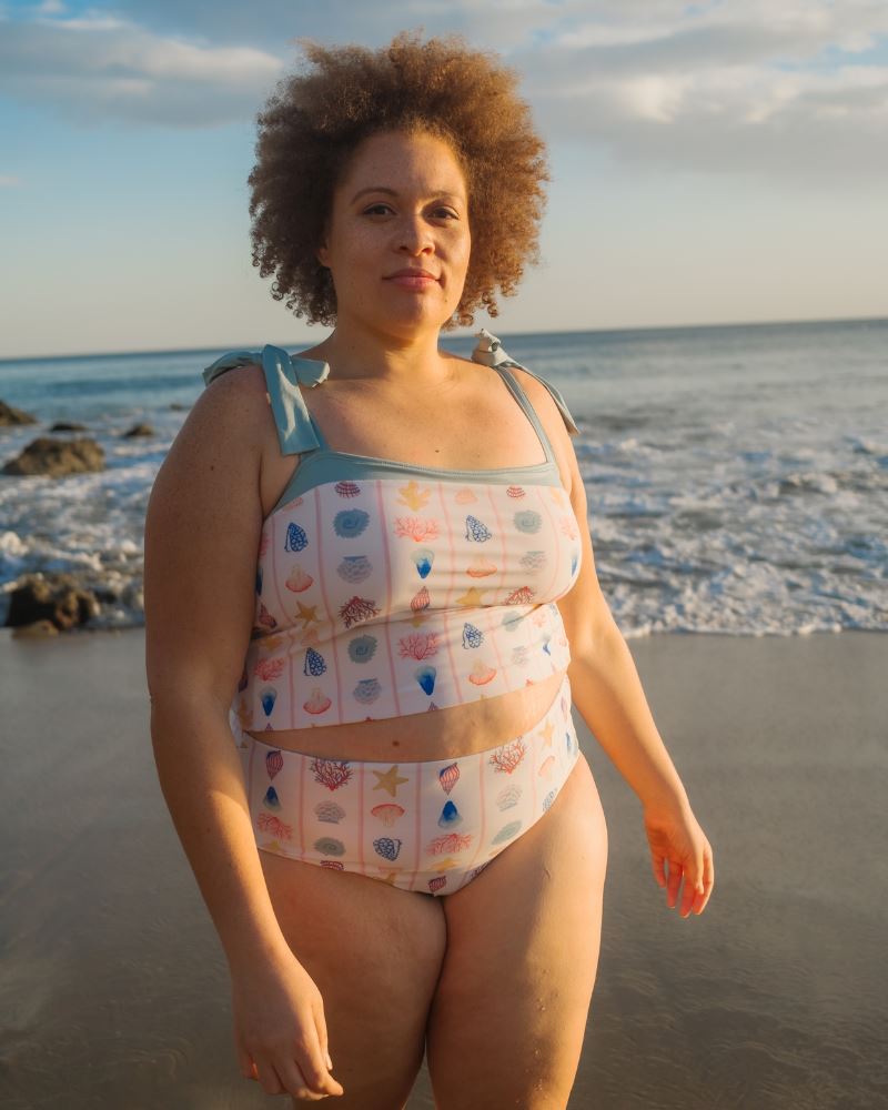 Photo of a woman posing on the beach wearing a seashell striped shoulder-tie swim crop top and a seashell striped high waist swim bottom