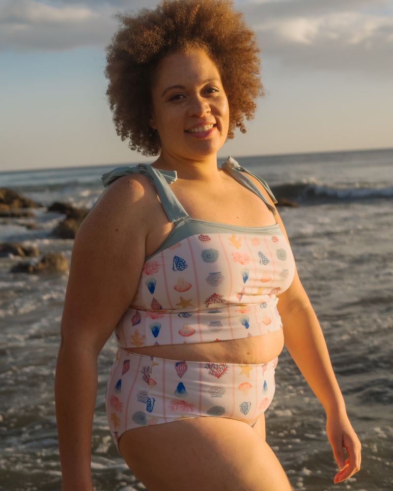 Photo of a woman posing on the beach wearing a seashell striped shoulder-tie swim crop top and a seashell striped high waist swim bottom