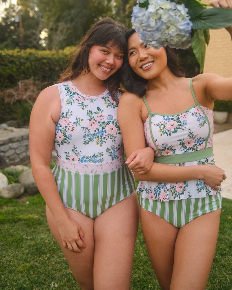Photo of two women standing together one wearing a pink and white floral one piece swimsuit the other wearing a pink and white floral swim top with green and white stripe high waist swim bottoms