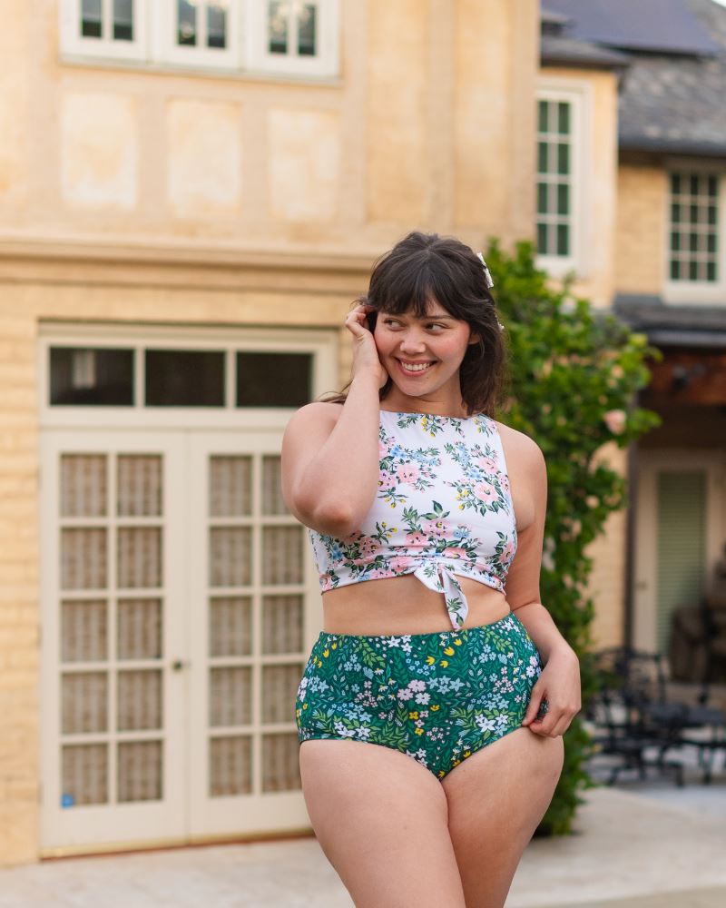 Photo of a woman posing outside wearing a dark green floral/ light green reversible swim bottom- floral side and a white and pink floral knotted swim crop top