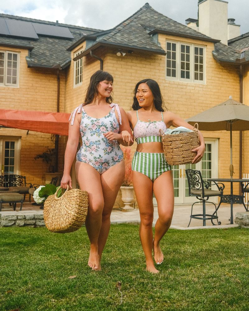 Photo of two women standing together one wearing a pink and white floral one piece swimsuit the other wearing a pink floral cropped swim top with green and white stripe high waist swim bottoms