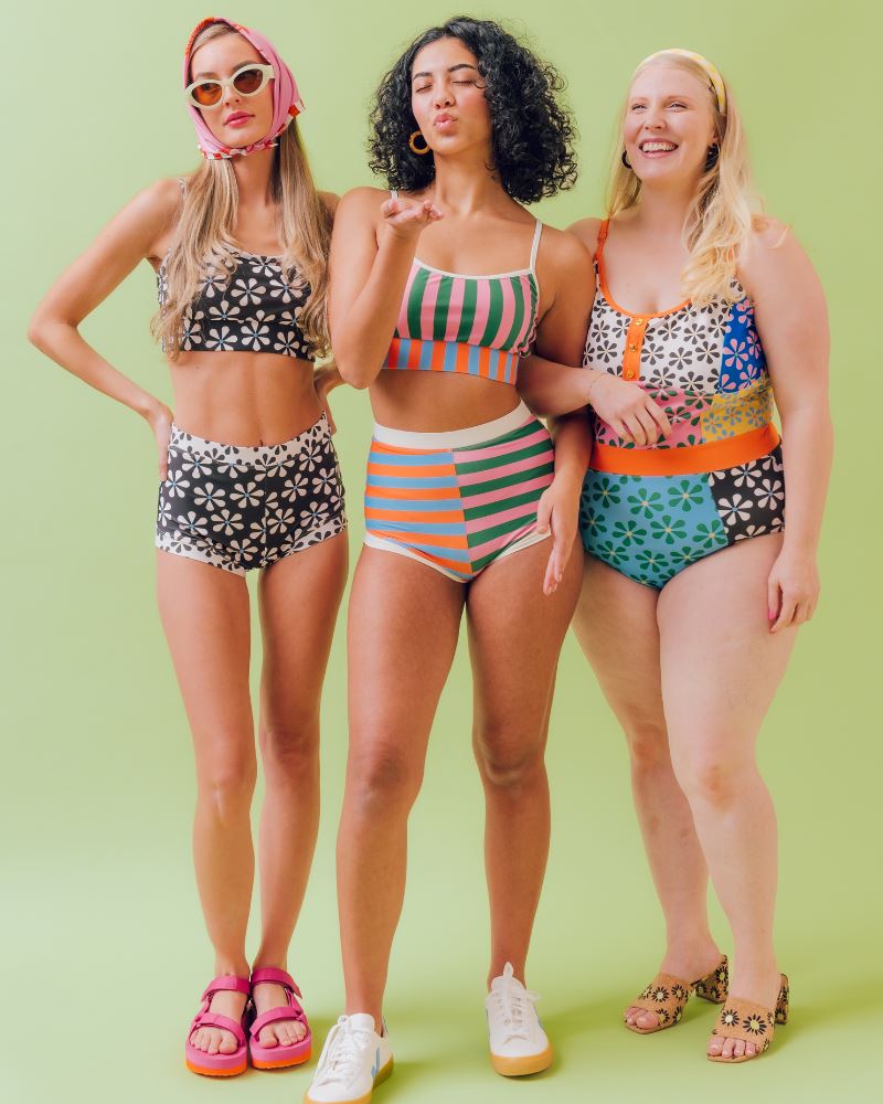 Photo of three women posing together (from left to right) wearing a black and white floral swim bralette and a black and white floral swim short bottom, another woman wearing a multi colored striped swim bralette and a multi colored striped swim short bottom, and another woman wearing a multi-colored floral one-piece swimsuit