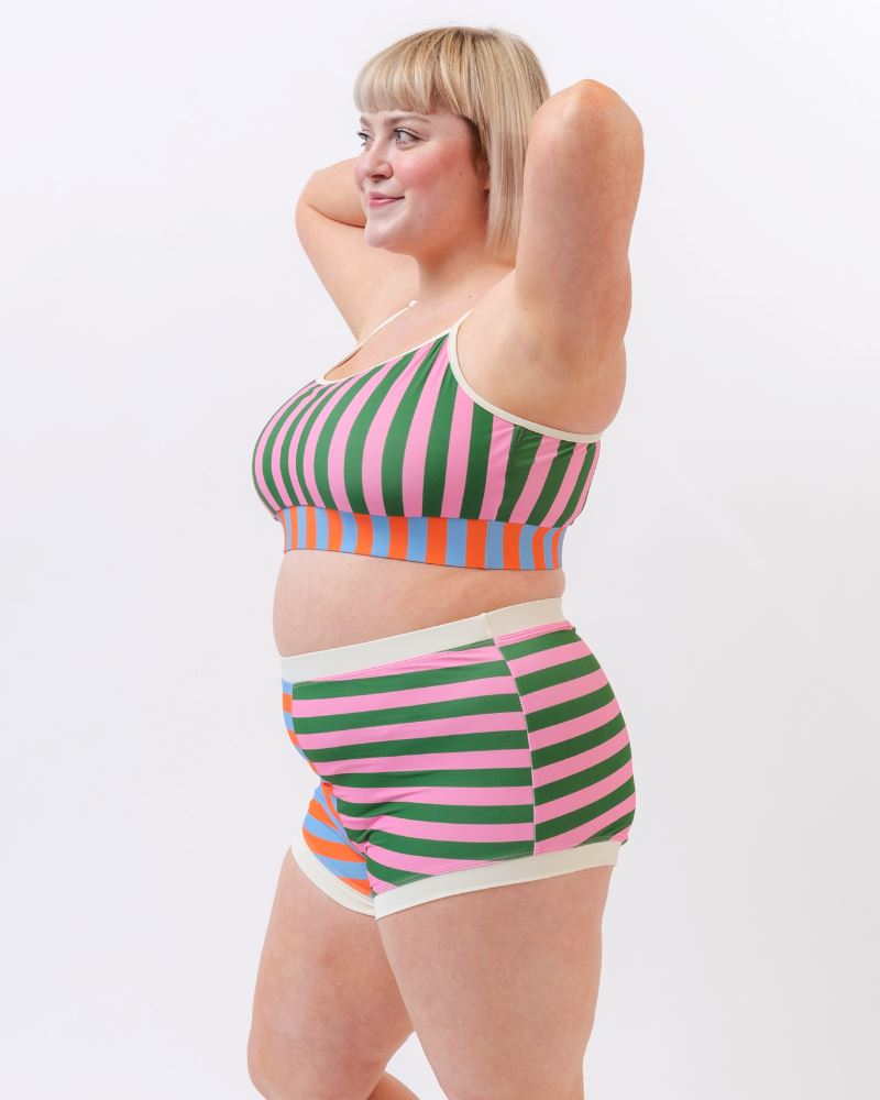 Photo of a woman wearing a multi-colored striped swim bralette and a multi-colored striped retro swim short bottom- side angle