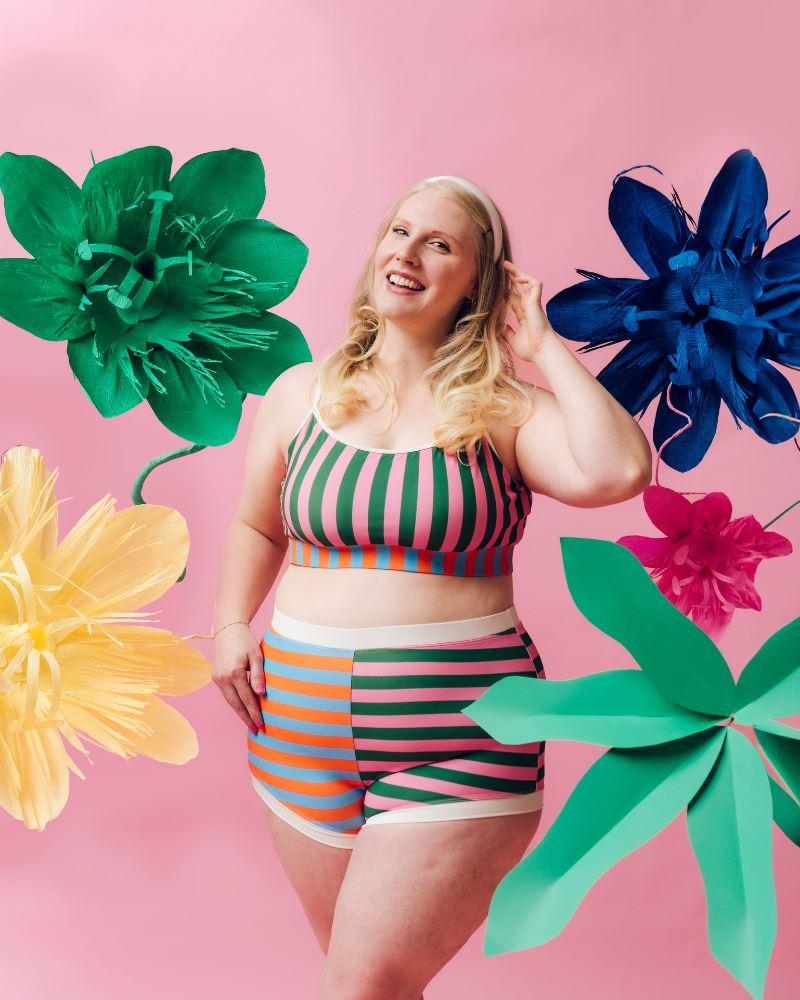 Photo of a woman posing with large flowers wearing a multi-colored striped swim bralette and a multi-colored striped retro swim short bottom