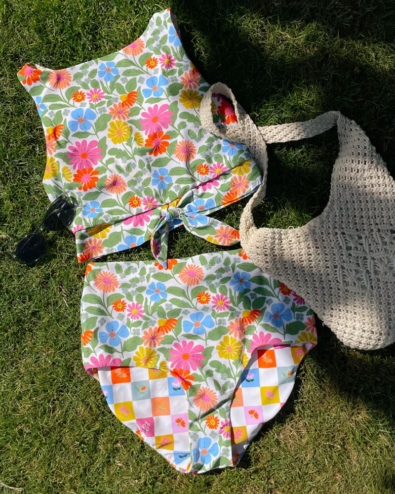 Photo of a multi color floral cropped swim top with multi color floral high waist swim bottoms laying in the grass with a beach bag and sunglasses