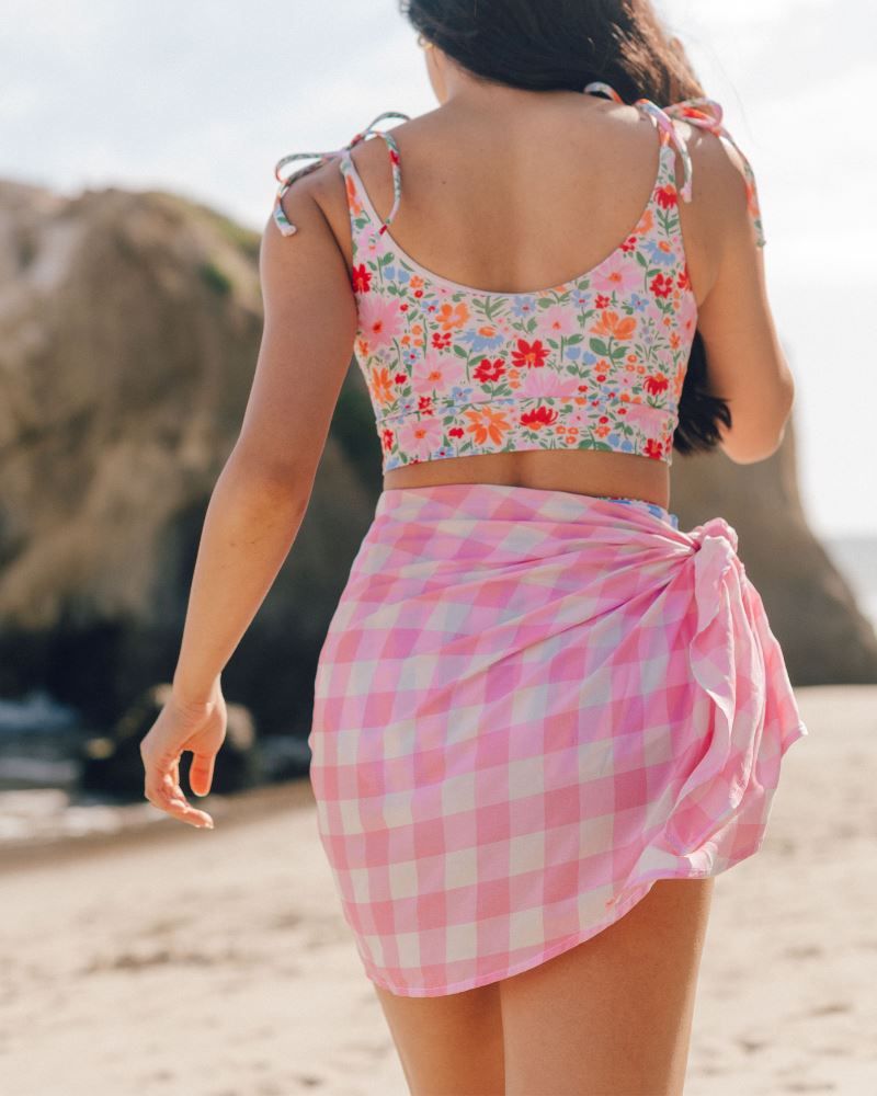 Photo of a woman wearing a pink and white checkered Sarong and a floral swim crop top back angle