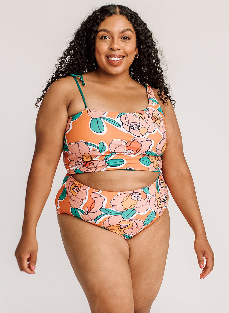 Photo of a woman wearing an orange and green floral one-shoulder swim crop top and an orange and green floral swim bottom