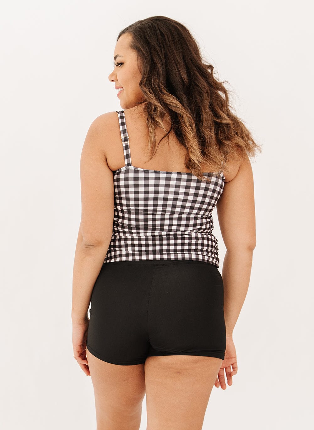Photo of a woman wearing high-waist black swim shorts and a black gingham square neck tankini swim top back angle