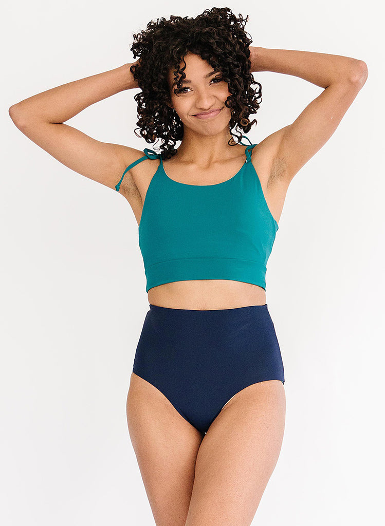 Photo of a woman with her hands in her hair wearing a blue cropped swim top with blue high waist swim bottoms