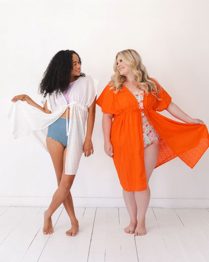 Photo of two women wearing an orange Kaftan cover up and floral swim suit and a white Kaftan Cover-Up and blue and white stripe swim bottom and a lilac swim crop top