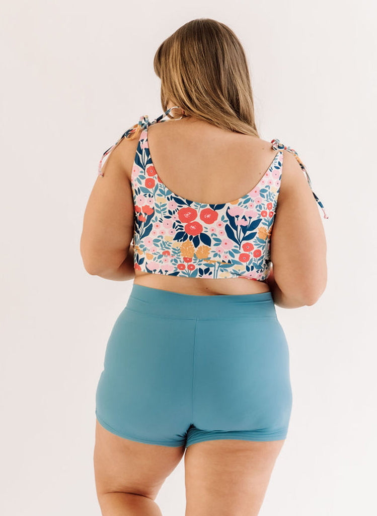 Photo of a woman wearing a may Flowers shoulder-tie swim crop top and an ocean blue swim short bottom back angle