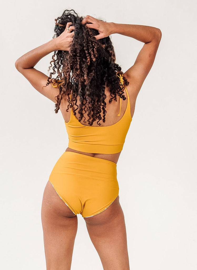 Photo of a woman with her back facing us with her hands in her hair while wearing a yellow cropped swim top with yellow high waist swim bottoms