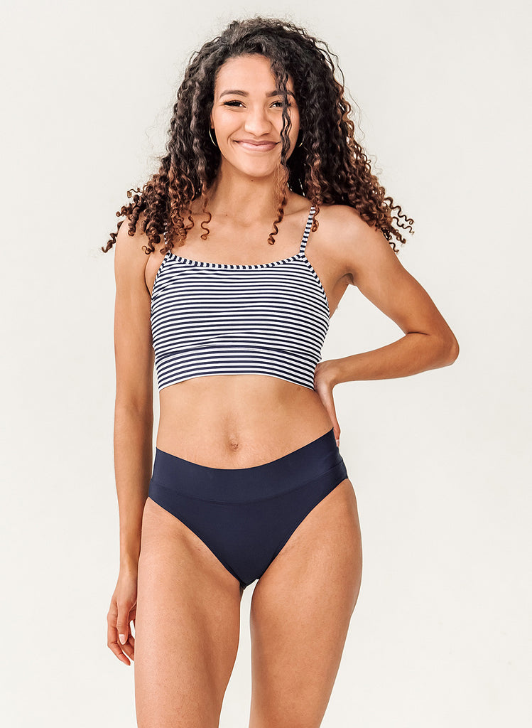 Photo of a woman with her hand on her hip wearing a blue and white striped cropped swim top with blue classic low rise swim bottoms