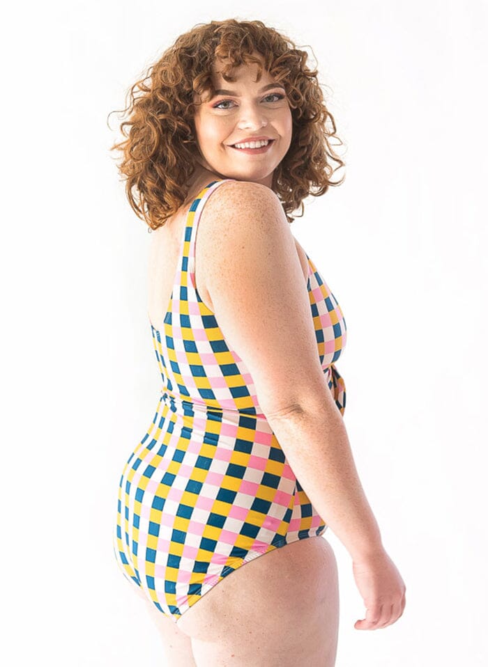 Photo of a woman with her back facing us looking over her shoulder while wearing a multi colored checkered one piece swim suit
