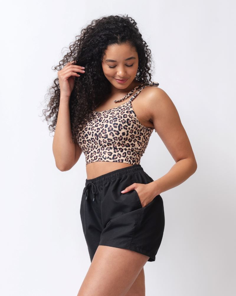 Photo of a woman wearing a black board short swim bottom and a leopard print shoulder-tie swim crop top- side angle
