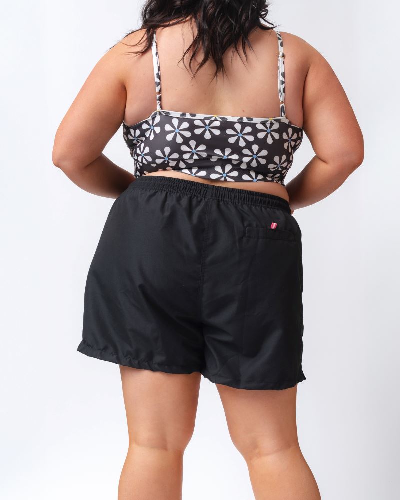 Photo of a woman wearing a black board short swim bottom and a black and white floral swim bralette- back angle