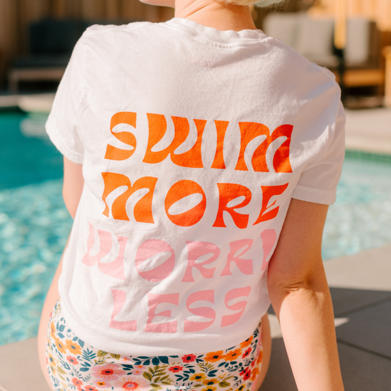Photo of woman sitting at pool with her back facing us wearing a Lime Ricki "Swim more, Worry Less" t-shirt with multi colored floral high waist swim bottoms