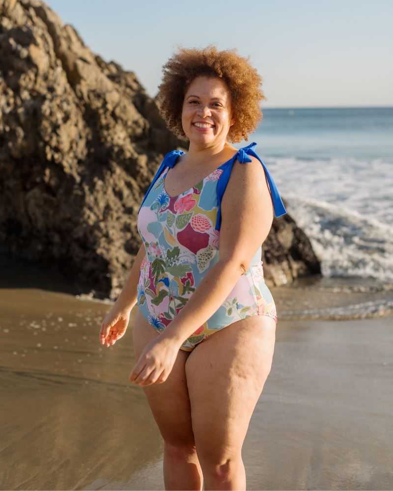 Photo of a woman posing on the beach wearing a colorful seashell inspired shoulder-tie one-piece swim suit- side angle