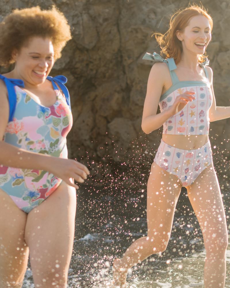 Photo of two women running together on the beach wearing a colorful seashell inspired shoulder-tie one-piece swim suit and the other woman wearing a seashell striped swim crop with a matching swim bottom