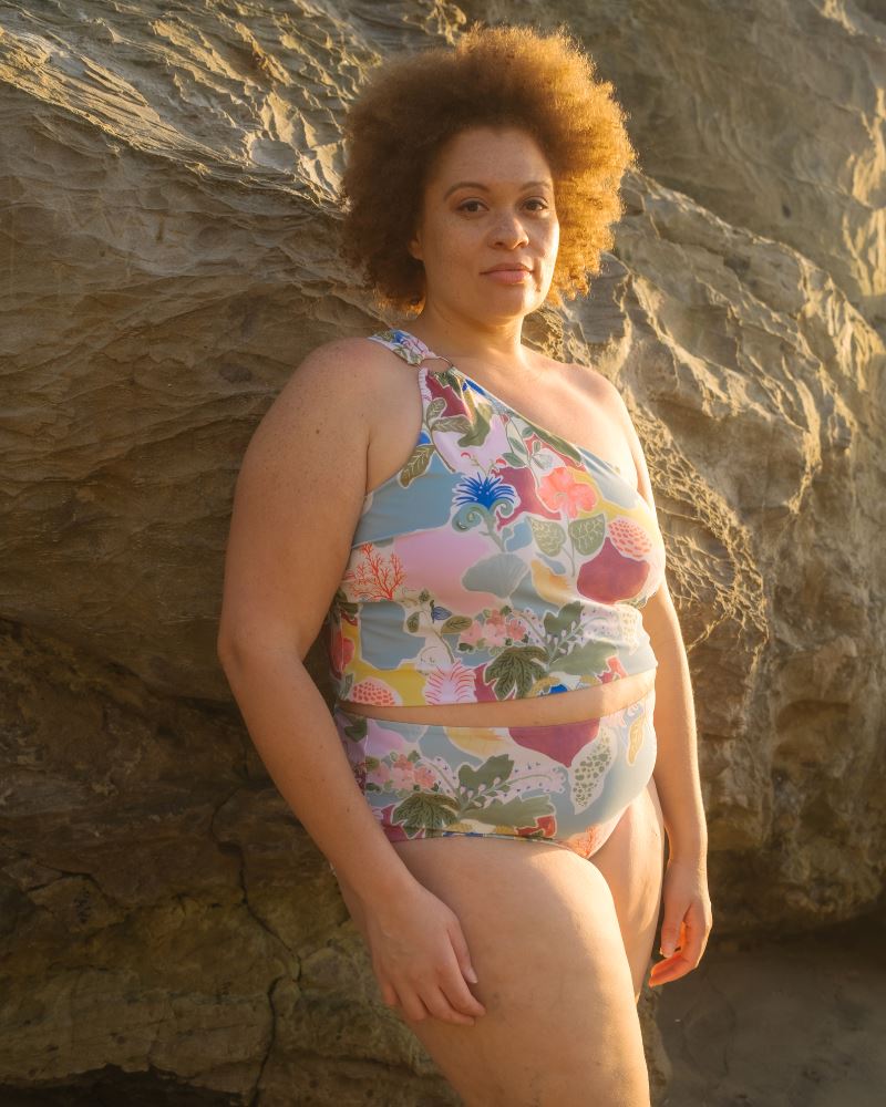 Photo of a woman posing on the beach wearing a colorful seashell inspired one-shoulder swim crop top and a colorful seashell inspired high waist swim bottom