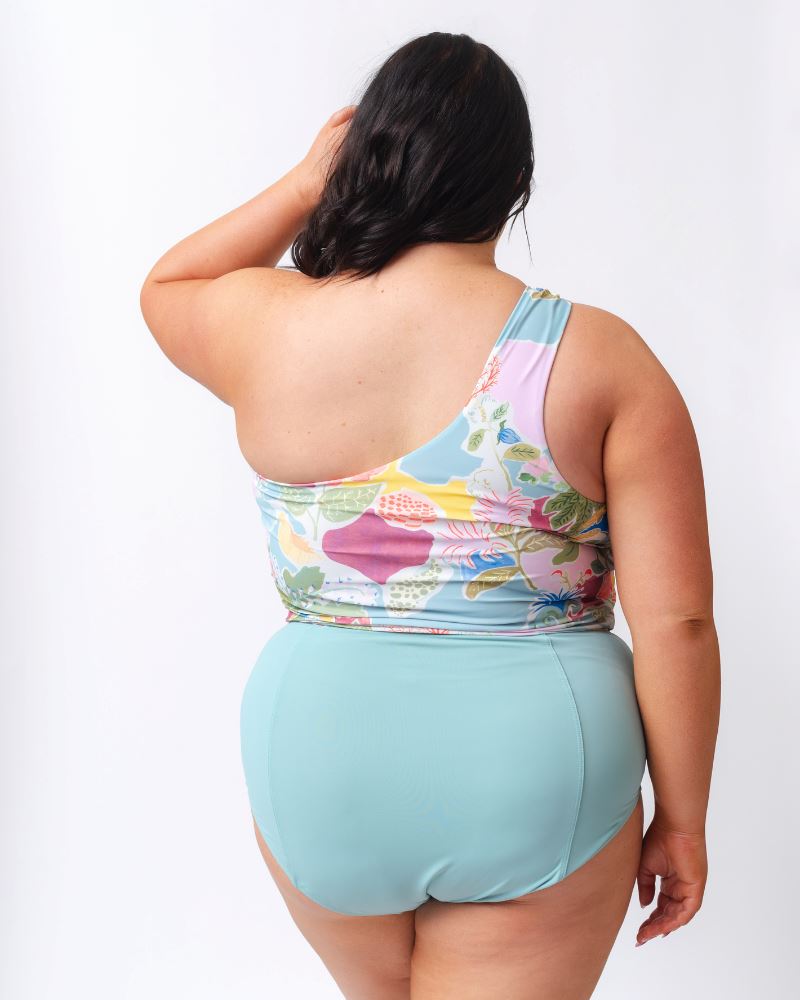 Photo of a woman wearing a light blue high waist swim bottom and a colorful seashell inspired one-shoulder swim crop top- back angle