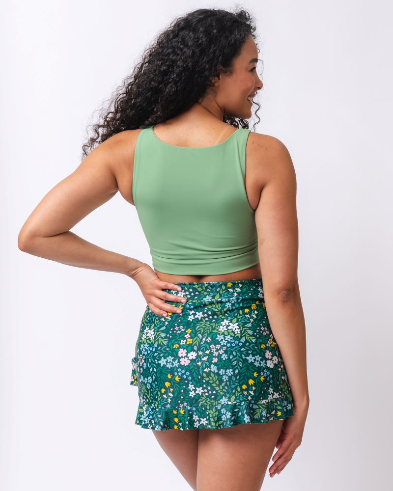 Photo of a woman wearing a dark green floral swim skirt and a ligh green knotted swim crop top- back angle