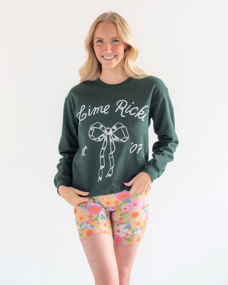 Photo of woman smiling while wearing a green Lime Ricki crew neck sweatshirt with multi colored floral long swim shorts